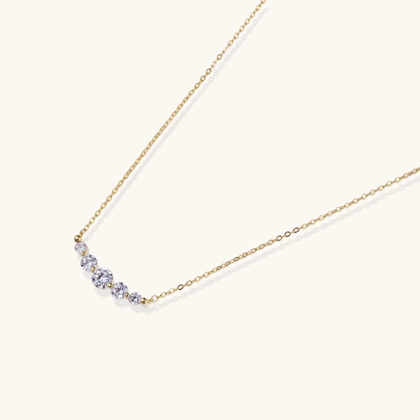 Delicate 14K gold plated necklace with curved arc of sparkling zirconia stones on cream background.