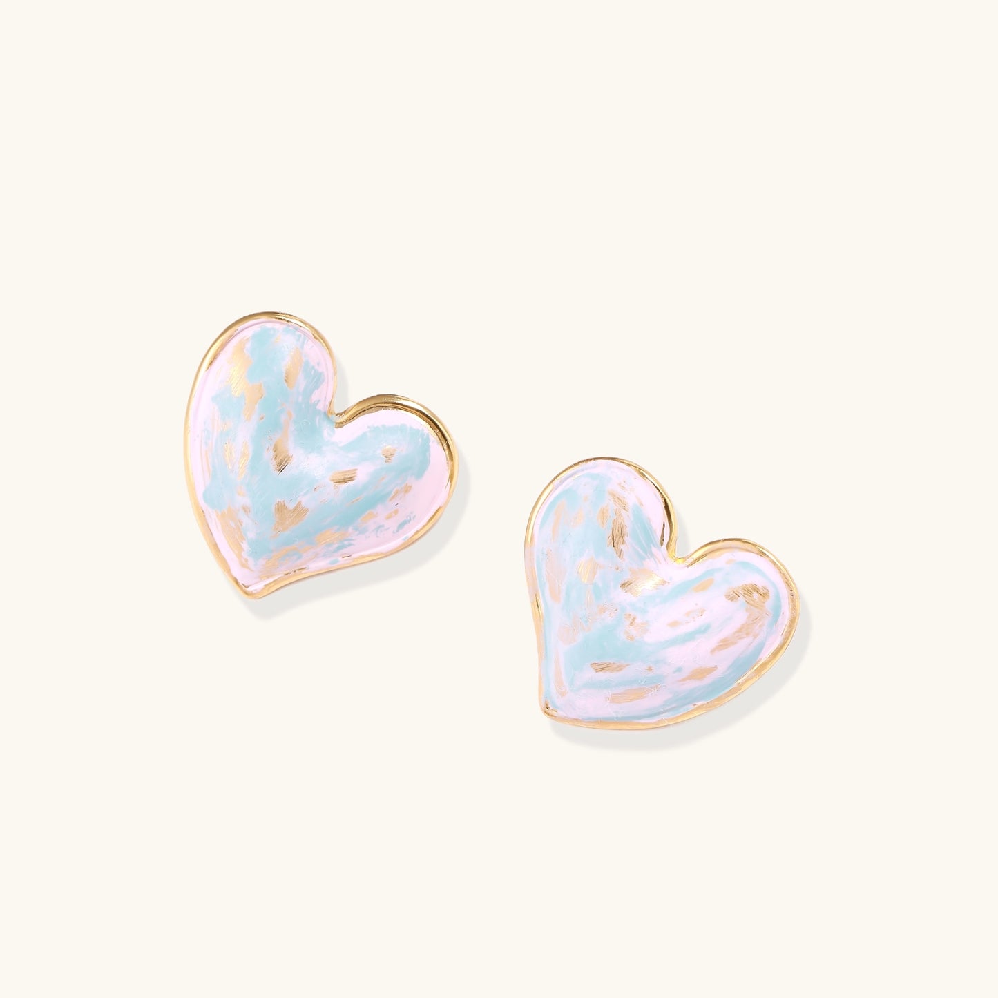 Heart-shaped stud earrings with iridescent blue-pink enamel finish and gold-plated brass frames against cream background