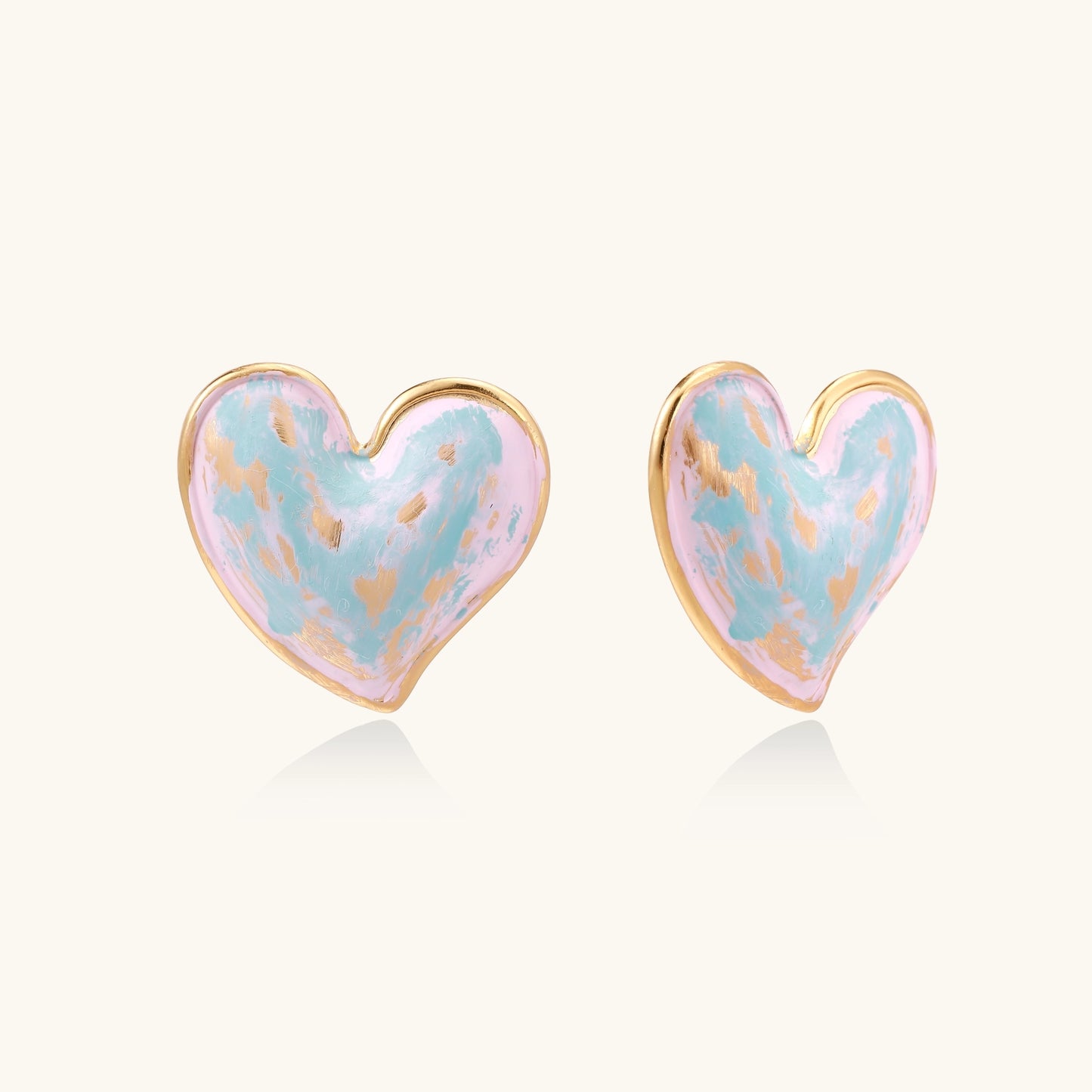 Heart-shaped stud earrings with iridescent blue-pink enamel finish and gold-plated brass frames against cream background